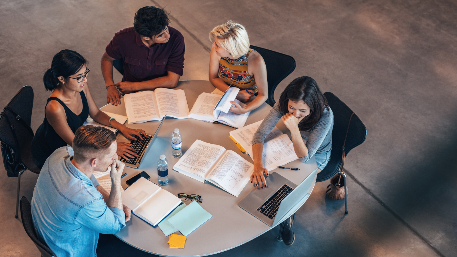 A group of students are seated at a circular table. There are books and an open laptop on the table